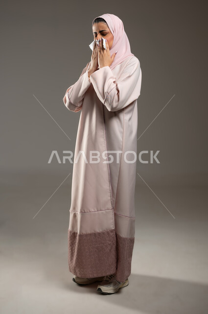 الاصابة بفايروس كورونا كوفيد19، استعمال المناديل الورقيه أثناء العطس، الشعور بالتعب والمرض، بورتريه من الجانب لامرأه عربيه خليجيه سعوديه محجبة ترتدي عباءة وردية، إيماءات وجه ويدين تدل على العطاس، صورة الجسم بالطول الكامل، خلفية رمادية، محتوى حساس، استخدام حساس