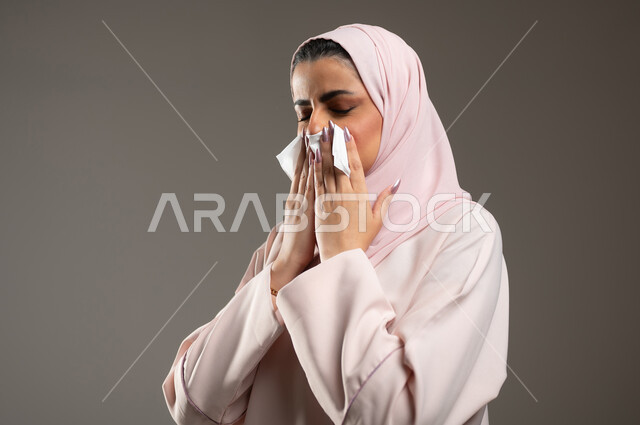 الشعور بالتعب والمرض، الإصابة بفايروس كورونا كوفيد19، استعمال المناديل الورقيه أثناء العطس، بورتريه من الجانب لامرأه عربيه خليجيه سعوديه محجبة ترتدي عباءة وردية، إيماءات وجه ويدين تدل على العطاس، خلفية رمادية، محتوى حساس، استخدام حساس