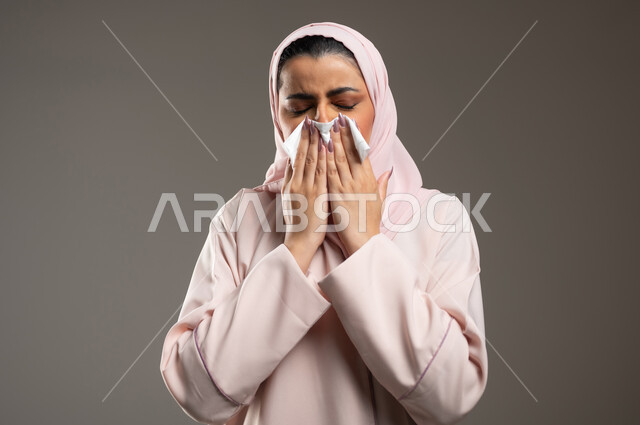 استعمال المناديل الورقية أثناء العطس، الشعور بالتعب والمرض، الإصابة بفايروس كورونا كوفيد19، بورتريه لامرأه عربيه خليجيه سعوديه محجبة ترتدي عباءة وردية، إيماءات وجه ويدين تدل على العطاس، خلفية رمادية، محتوى حساس، استخدام حساس
