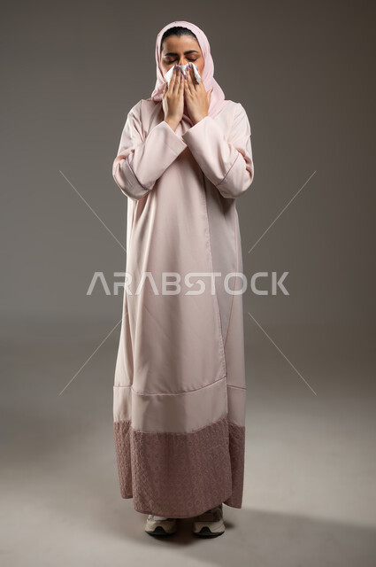 فايروس كورونا كوفيد19، استعمال المناديل الورقيه أثناء العطس، الشعور بالتعب والمرض، بورتريه لامرأه عربيه خليجيه سعوديه محجبة ترتدي عباءة وردية، إيماءات وجه ويدين تدل على العطاس، صورة الجسم بالطول الكامل، خلفية رمادية، محتوى حساس، استخدام حساس