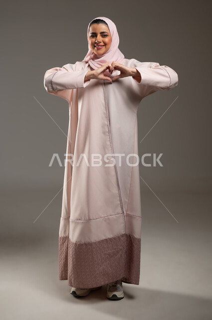ايماءات يدين على شكل قلب، اظهار مشاعر الحب والشغف، مفهوم المحبة والمودة، بورتريه لامرأه عربيه خليجيه سعوديه محجبة ترتدي عباءة وردية تنظر إلى الكاميرا بتعابير السرور والسعادة، صورة الجسم بالطول الكامل، خلفية رمادية، محتوى حساس، استخدام حساس