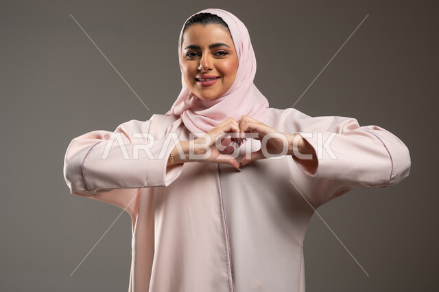 مفهوم المحبة والمودة، اظهار مشاعر الحب والشغف، ايماءات يدين على شكل قلب، بورتريه مقرب لامرأه عربيه خليجيه سعوديه محجبة ترتدي عباءة وردية تنظر إلى الكاميرا بتعابير السرور والسعادة، خلفية رمادية، محتوى حساس، استخدام حساس
