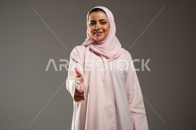 Expressions of contentment and pleasure, feeling of positivity and happiness, the concept of greetings and peace, a close-up portrait of a veiled Saudi Gulf Arab woman wearing a pink abaya with hand gestures indicating a handshake, gray background, sensitive content, sensitive use