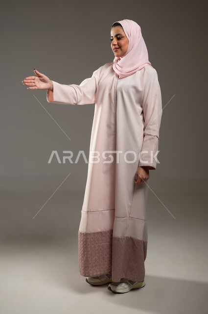 الشعور بالإيجابيه والسعادة، مفهوم التحيه والسلام، بورتريه لامرأه عربيه خليجيه سعوديه محجبة ترتدي عباءة وردية بإيماءات يد تدل على المصافحه، تعابير الرضى والسرور، صورة الجسم بالطول الكامل، خلفية رمادية، محتوى حساس، استخدام حساس