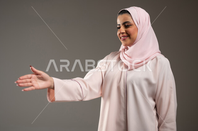 مفهوم التحيه والسلام، بورتريه مقرب لامرأه عربيه خليجيه سعوديه محجبة ترتدي عباءة وردية بإيماءات يد تدل على المصافحه، الشعور بالإيجابيه والسعادة، تعابير الرضى والسرور، خلفية رمادية، محتوى حساس، استخدام حساس