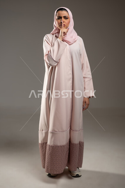 الأمر بالصمت والتوقف عن الكلام، مفهوم عدم الإزعاج، بورتريه من الجانب لامرأه عربيه خليجيه سعودية محجبة ترتدي عباءة وردية إيماءات يدين تدل على طلب السكوت والهدوء، صورة الجسم بالطول الكامل، خلفية رمادية، محتوى حساس، استخدام حساس