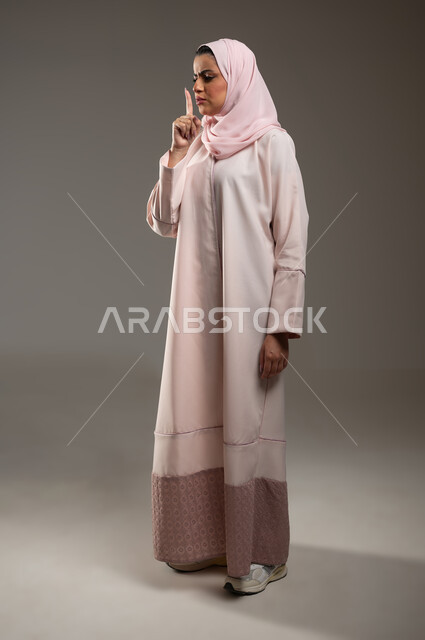 The concept of not disturbing, a side portrait of a veiled Saudi Gulf Arab woman wearing a pink abaya, two hand gestures indicating a request for silence and calm, the command to be silent and stop talking, full-length body image, gray background, sensitive content, sensitive use