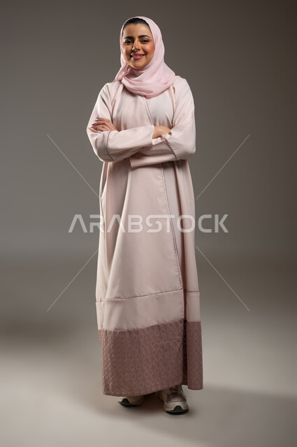 Gestures of self-confidence, attention to elegance and external appearance, the concept of femininity and softness, a portrait of a veiled Saudi Gulf Arab woman wearing a pink abaya looking at the camera with an expression of pleasure, full-length body image, gray background, sensitive content, sensitive use