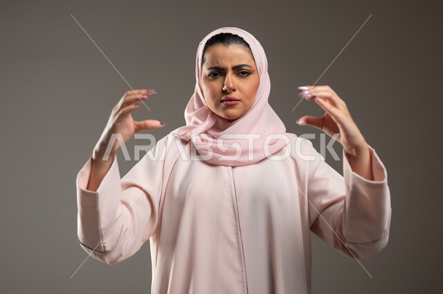 ايماءات الغضب والعصبية، تعابير الجدية والانزعاج، بورتريه مقرب لامرأة عربية خليجية سعودية محجبة ترتدي عباءة وردية تمسك بعنصر ما، خلفية رمادية، محتوى حساس، استخدام حساس