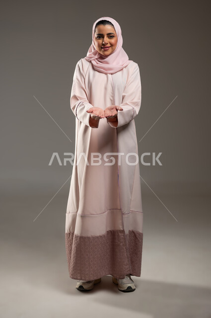 النظر إلى الكاميرا بايماءات السعادة والسرور، بورتريه لامرأة عربية خليجية سعودية محجبة ترتدي عباءة وردية تفتح يديها لعرض شيء ما، حمل ورفع عنصر ما على الكفين، صورة الجسم بالطول الكامل، خلفية رمادية، محتوى حساس، استخدام حساس