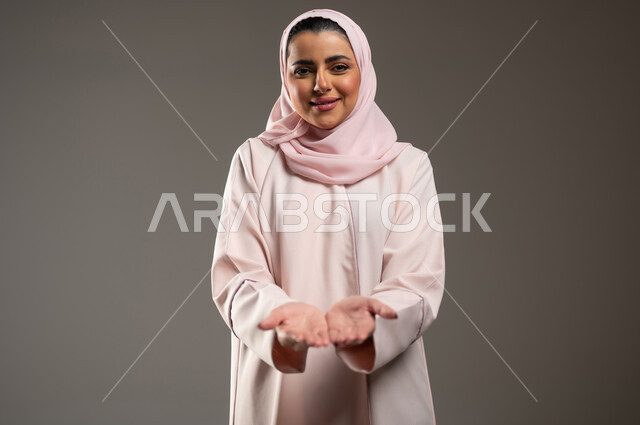 حمل ورفع عنصر ما على الكفين، النظر إلى الكاميرا بايماءات السعادة والسرور، بورتريه مقرب لامرأة عربية خليجية سعودية محجبة ترتدي عباءة وردية تفتح يديها لعرض شيء ما، خلفية رمادية، محتوى حساس، استخدام حساس