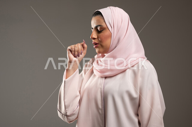 الشعور بالمرض والكحة، بورتريه مقرب من الجانب لامرأه عربية خليجية سعودي محجبة ترتدي عباءة وردية تعاني من السعال، الانفلونزا والتهاب الرئه، الاصابة بفايروس كورونا كوفيد19، خلفية رمادية، محتوى حساس، استخدام حساس