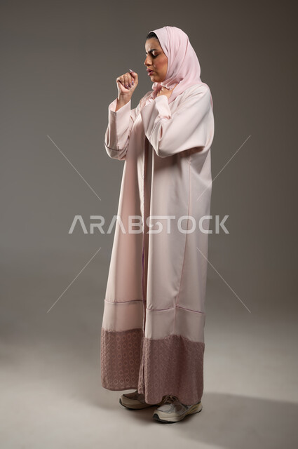 Flu and pneumonia, feeling sick and coughing, portrait from the side of a veiled Saudi Gulf Arab woman wearing a pink abaya suffering from cough, infection with the Corona virus Covid-19, full-length body image, gray background, sensitive content, sensitive use