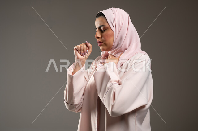 الشعور بالمرض والكحة، بورتريه مقرب من الجانب لامرأه عربية خليجية سعودي محجبة ترتدي عباءة وردية تعاني من السعال، الانفلونزا والتهاب الرئه، الاصابة بفايروس كورونا كوفيد19، خلفية رمادية، محتوى حساس، استخدام حساس