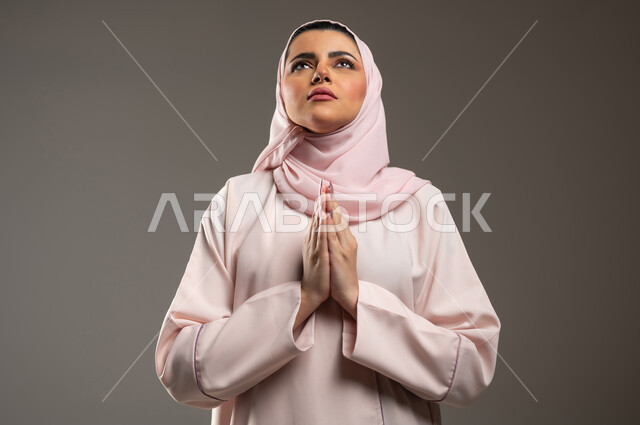 طلب المغفره من الله، الرغبه في تحقيق الأمنيات، مفهوم طلب العون، بورتريه مقرب لامرأة عربية خليجية سعودية محجبة ترتدي عباءة وردية ترفع يديها بايماءات تدل على الدعاء والتضرع، خلفية رمادية، محتوى حساس، استخدام حساس