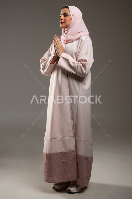 مفهوم طلب العون، الحمد والشكر لله، بورتريه من الجانب لامرأة عربية خليجية سعودية محجبة ترتدي عباءة وردية ترفع يديها بايماءات تدل على الدعاء والتضرع، الرغبه في تحقيق الأمنيات، صورة الجسم بالطول الكامل، خلفية رمادية، محتوى حساس، استخدام حساس