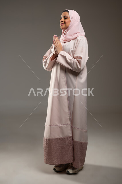 مفهوم طلب العون، الحمد والشكر لله، بورتريه من الجانب لامرأة عربية خليجية سعودية محجبة ترتدي عباءة وردية ترفع يديها بايماءات تدل على الدعاء والتضرع، الرغبه في تحقيق الأمنيات، صورة الجسم بالطول الكامل، خلفية رمادية، محتوى حساس، استخدام حساس