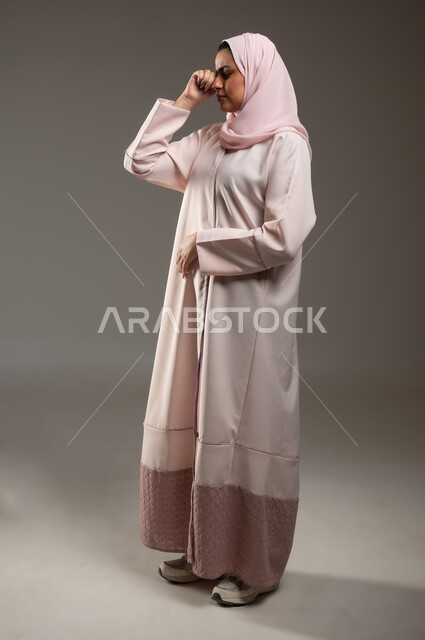 الندم والحسرة على شيء ما، مفهوم القهر والخسارة، بورتريه من الجانب لامرأه عربيه خليجيه سعوديه محجبة ترتدي عباءة وردية تضع يدها على عينها بإيماءات تدل على البكاء والحزن، الشعور بالإنزعاج والخذلان، صورة الجسم بالطول الكامل، خلفية رمادية، محتوى حساس، استخدام حساس