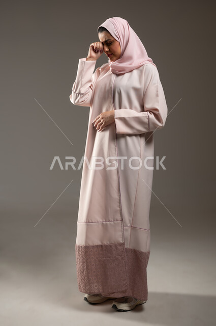 مفهوم القهر والخسارة، الندم والحسرة على شيء ما، بورتريه من الجانب لامرأه عربيه خليجيه سعوديه محجبة ترتدي عباءة وردية تضع يدها على عينها بإيماءات تدل على البكاء والحزن، الشعور بالإنزعاج والخذلان، صورة الجسم بالطول الكامل، خلفية رمادية، محتوى حساس، استخدام حساس