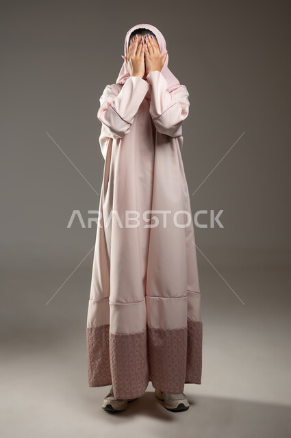 إغماض العينين وانتظار رؤية المفاجأة، بورتريه لامرأه عربيه خليجيه سعوديه محجبة ترتدي عباءة وردية تضع كفيها على وجهها بإيماءات تدل على البكاء والحزن، صورة الجسم بالطول الكامل، خلفية رمادية، محتوى حساس، استخدام حساس