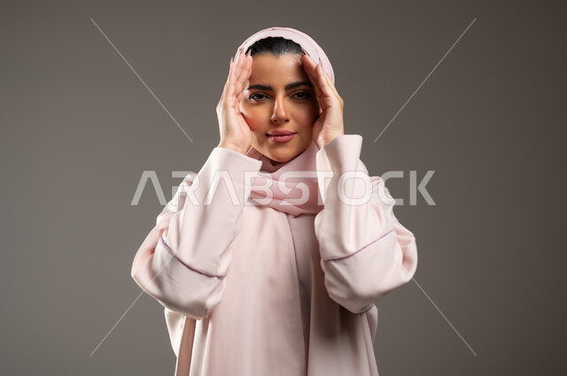Gestures of pleasure and happiness, focusing and looking at something, close-up portrait of a veiled Saudi Gulf Arab woman wearing a pink abaya, placing her palms on her face with expressions indicating surveillance, gray background, sensitive content, sensitive use