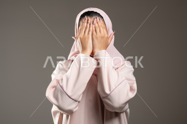Gestures of closing the eyes and waiting to see the surprise, a close-up portrait of a veiled Saudi Gulf Arab woman wearing a pink abaya, placing her palms on her face with expressions indicating crying and sadness, gray background, sensitive content, sensitive use