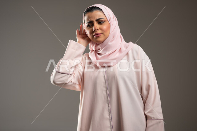 استراق السمع والتجسس، تعابير التركيز والاستماع، بورتريه مقرب لإمرأة عربية خليجية سعودية محجبة ترتدي عباءة وردية تضع يدها على اذنها بايماءات الاصغاء، خلفية رمادية، محتوى حساس، استخدام حساس