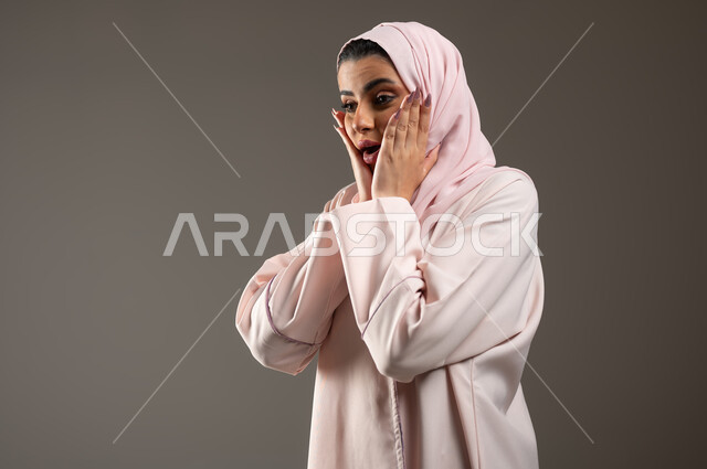 النظر إلى مكان ما بإيماءات الدهشة والاستغراب، التعجب من حصول أمر ما، بورتربه مقرب من الجانب لامرأة عربية خليجية سعودية محجبة ترتدي عباءة وردية تضع يديها على وجهها بإيماءات تدل على الصدمة والذهول، خلفية رمادية، محتوى حساس، استخدام حساس