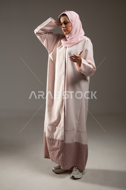 الشعور بالحزن والضجر، الانزعاج لسماع الأخبار السيئة، بورتريه من الجانب لإمرأة عربية خليجية سعودية محجبة ترتدي عباءة وردية تضع يدها على رأسها وترفع الاخرى بايماءات التساؤل، صورة الجسم بالطول الكامل، خلفية رمادية، محتوى حساس، استخدام حساس