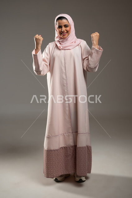 تحقيق الأهداف والنجاح، مفهوم الفرح بالنصر والفوز، تعابير السرور والسعادة، بورتريه لامرأة عربية خليجية سعودية محجبة مبتسمة ترتدي عباءة وردية ترفع يديها بإيماءات الحماس، صورة الجسم بالطول الكامل، خلفية رمادية، محتوى حساس، استخدام حساس