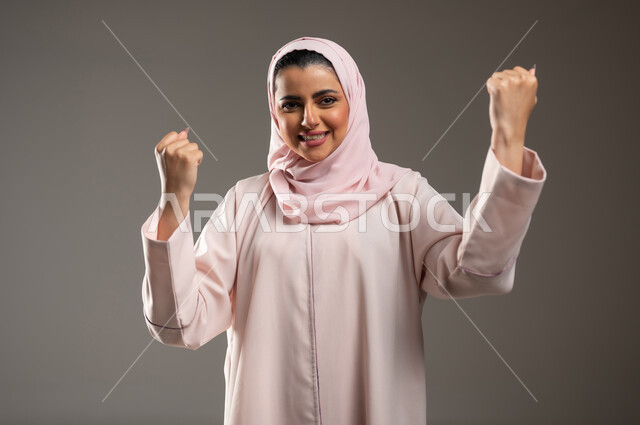 النجاح وتحقيق الأهداف، مفهوم الفرح بالنصر والفوز، تعابير السرور والسعادة، بورتريه مقرب لامرأة عربية خليجية سعودية محجبة مبتسمة ترتدي عباءة وردية ترفع يديها بإيماءات الحماس، خلفية رمادية، محتوى حساس، استخدام حساس