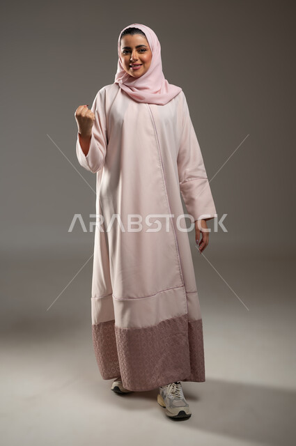 تحقيق الأهداف والنجاح، تعابير السرور والسعادة، مفهوم الفرح بالنصر والفوز، بورتريه لامرأة عربية خليجية سعودية محجبة مبتسمة ترتدي عباءة وردية ترفع يدها بإيماءات الحماس، صورة الجسم بالطول الكامل، خلفية رمادية، محتوى حساس، استخدام حساس