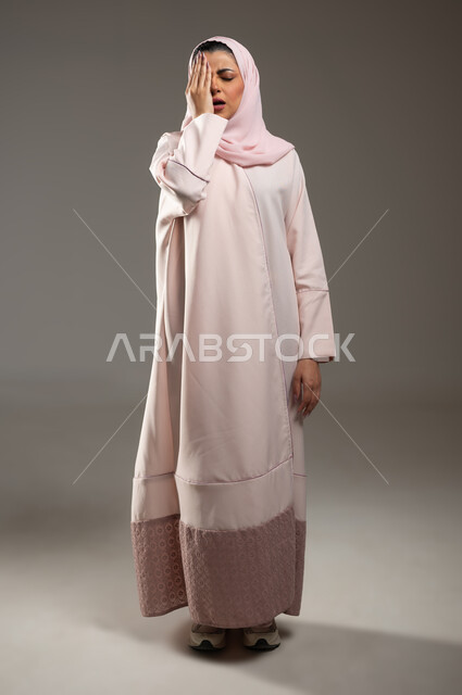 Migraine pain and migraine, the need for calm and rest, the feeling of headache and headache, a portrait of a veiled Saudi Arabian Gulf woman wearing a pink abaya, placing her hand on her face with gestures of fatigue and exhaustion, full-length body image, gray background, sensitive content, sensitive use