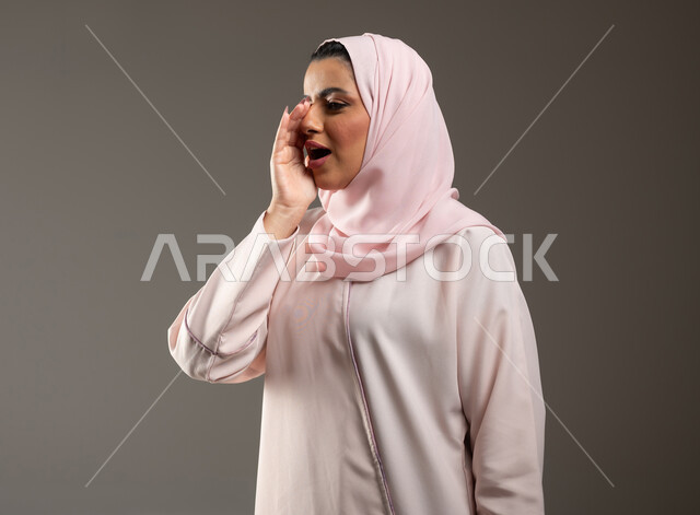 مفهوم الدردشات والمحادثات، النداء بصوت منخفض والكلام بصوت عالي، بورتريه مقرب لامرأة عربية خليجية سعودية محجبة ترتدي عباءة وردية تضع يدها على فمها بإيماءات تدل على المناداة، خلفية رمادية، محتوى حساس، استخدام حساس