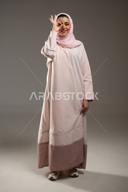 مفهوم الرؤية المستقبلية، حب البحث والاستكشاف والمغامرة، بورتريه لامرأة عربية خليجية سعودية محجبة مبتسمة ترتدي عباءة وردية تضع يدها على عينها بشكل عدسة بإيماءات و تعابير وجه ويدين تدل على المشاهدة، صورة لكامل الجسم، خلفية رمادية، محتوى حساس، استخدام حساس