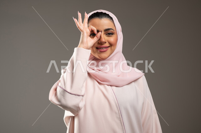 حب البحث والاستكشاف والمغامرة، بورتريه مقرب لامرأة عربية خليجية سعودية محجبة مبتسمة ترتدي عباءة وردية تضع يدها على عينها بشكل عدسة بإيماءات و تعابير وجه ويدين تدل على المشاهدة، مفهوم الرؤية المستقبلية، خلفية رمادية، محتوى حساس، استخدام حساس