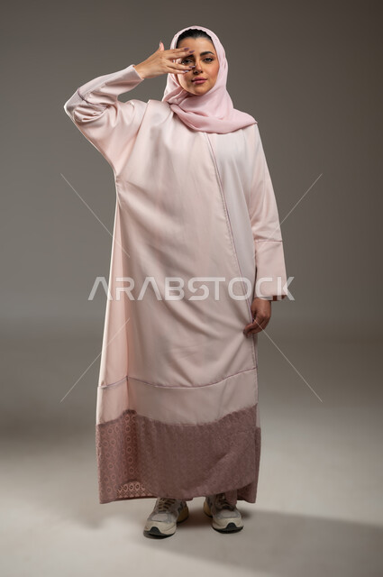التحديق الى شيء ما، بورتريه لامرأة عربية خليجية سعودية محجبة ترتدي عباءة وردية تضع اصابع يدها على عينها، النظر الى الكاميرا بايماءات التركيز، صورة الجسم بالطول الكامل، خلفية رمادية، محتوى حساس، استخدام حساس
