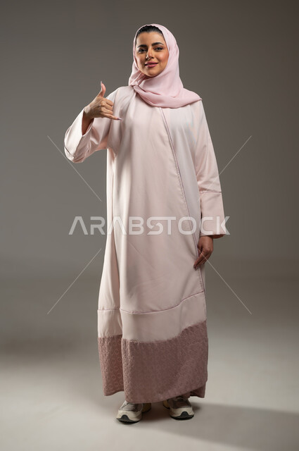 اجراء مكالمه هاتفيه، تعابير السعادة والسرور، بورتريه لامرأة عربية خليجية سعودية محجبة ترتدي عباءة وردية ترفع يدها بإيماءات الاتصال، مفهوم الدردشات والمحادثات، صورة الجسم بالطول الكامل، خلفية رمادية، محتوى حساس، استخدام حساس