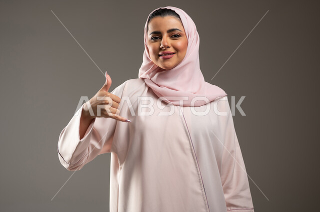 مفهوم الدردشات والمحادثات، تعابير السعادة والسرور، بورتريه مقرب لامرأة عربية خليجية سعودية محجبة ترتدي عباءة وردية ترفع يدها بإيماءات الاتصال، اجراء مكالمه هاتفيه، خلفية رمادية، محتوى حساس، استخدام حساس