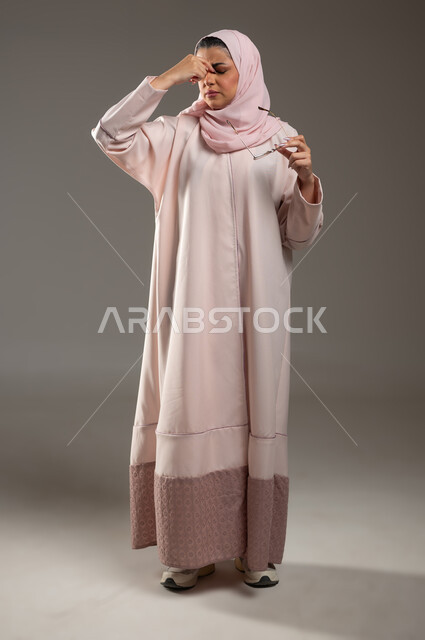 الشعور بالمرض والإرهاق، ضعف القدرة على الرؤية، بورتريه لامرأة عربية خليجية سعودية محجبة ترتدي عباءة وردية تمسك نظارة طبية تضع يدها بين عينيها بايماءات التعب، صورة لكامل الجسم، خلفية رمادية، محتوى حساس، استخدام حساس