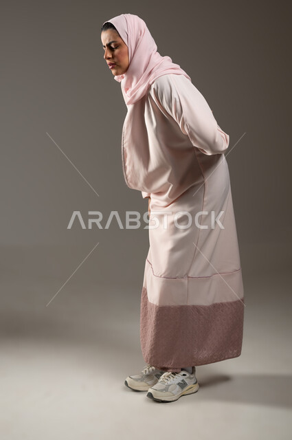 تعابير الألم ووجع الظهر، الشعور بالمرض والإرهاق، بورتريه لامرأة عربية خليجية سعودية محجبة ترتدي عباءة وردية تضع يدها على ظهرها بايماءات التعب، الحاجة للعلاج والاستشارة الطبية، صورة لكامل الجسم، خلفية رمادية، محتوى حساس، استخدام حساس