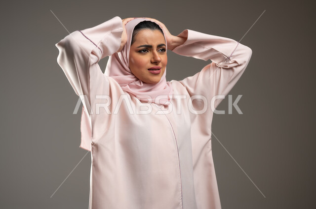 الانزعاج لسماع الأخبار السيئة، الشعور بالحزن والضجر، بورتريه مقرب لإمرأة عربية خليجية سعودية محجبة ترتدي عباءة وردية تضع يديها على رأسها تنظر إلى مكان ما بايماءات الصدمة، خلفية رمادية، محتوى حساس، استخدام حساس