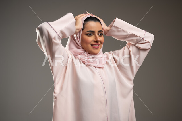 سماع الاخبار والمفاجأت السعيدة، ايماءات الدهشة والسرور، بورتريه مقرب لإمرأة عربية خليجية سعودية محجبة مبتسمة ترتدي عباءة وردية تضع يديها على رأسها تنظر إلى مكان ما بفرح وسعادة، خلفية رمادية، محتوى حساس، استخدام حساس