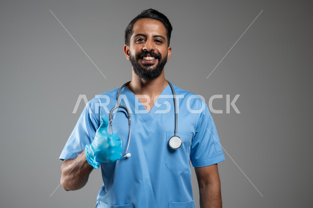النظر الى الكاميرا بإيماءات الموافقة والسرور, بورتريه مقرب لممرض عربي خليجي سعودي يرتدي القفازات واليونيفورم يرفع اصبع الإبهام للأعلى بتعابير علامة الجودة, الشعور بالرضا و القبول, خلفية رمادية, محتوى حساس, استخدام حساس