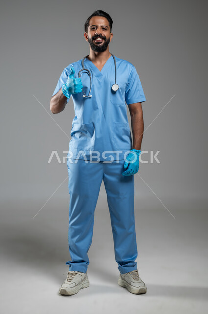 الشعور بالرضا و القبول, النظر الى الكاميرا بإيماءات الموافقة والسرور, بورتريه لممرض عربي خليجي سعودي يرتدي القفازات واليونيفورم يرفع اصبع الإبهام للأعلى بتعابير علامة الجودة, الجسم بالطول الكامل, خلفية رمادية, محتوى حساس, استخدام حساس