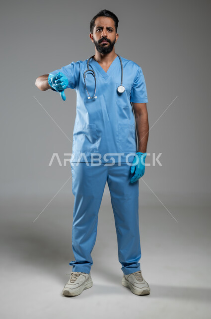 العمل في القطاع الصحي, تعابير الانزعاج وعدم القبول والرضا, مهن ووظائف طبية سعودية, بورتريه لممرض عربي خليجي يرتدي اليونيفورم الطبي يشير بإصبع الإبهام للأسفل بإيماءات الرفض وعدم الموافقة, صورة الجسم بالطول الكامل, خلفية رمادية, محتوى حساس, استخدام حساس