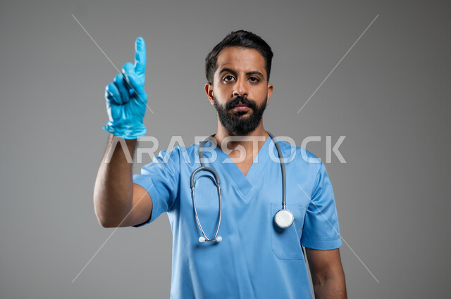 اشارة تمثل الرقم واحد, العمل في القطاع الصحي, بورتريه مقرب لممرض عربي خليجي سعودي يرتدي اليونيفورم الخاص بالعمل يرفع اصبع السبابة إلى الأعلى, النظر الى الكاميرا و الاشارة الى شيء ما, خلفية رمادية, محتوى حساس, استخدام حساس