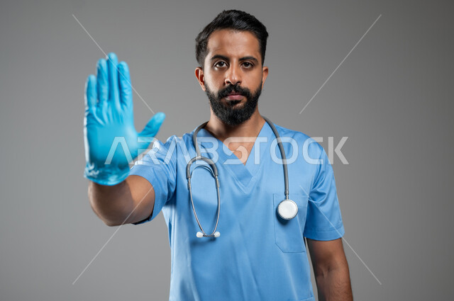 النظر إلى الكاميرا بايماءت الحزم والجدية, مهن ووظائف طبية سعودية, تقديم الخدمات الصحية للمرضى، بورتريه مقرب لممرض عربي خليجي سعودي يرتدي اليونيفورم الخاص بالعمل يرفع يده بتعابير طلب التوقف والابتعاد, خلفية رمادية, محتوى حساس, استخدام حساس