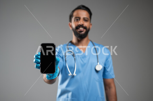 مفهوم الطب والرعاية الصحية, دمج التقنيات الحديثة في الحياة العملية, بورتريه مقرب لممرض عربي سعودي خليجي يرتدي يونيفورم العمل والقفازات يقوم بعرض شاشة سوداء فارغه عبر الهاتف المحمول، مهن ووظائف سعودية, خلفية رمادية، محتوى حساس، استخدام حساس