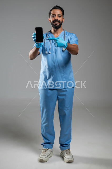 النظر الى الكاميرا بتعابير السرور, العمل في القطاع الصحي, دمج التكنولوجيا في الحياة العملية, بورتريه لطبيب عربي سعودي خليجي يرتدي يونيفورم العمل يشير باصبع السبابة الى شاشة سوداء فارغة عبر الجوال، الطول الكامل للجسم, خلفية رمادية، محتوى حساس، استخدام حساس